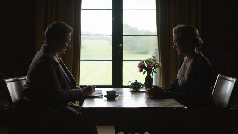 Two women in period costume face each other across a table, silhouetted against a tall window